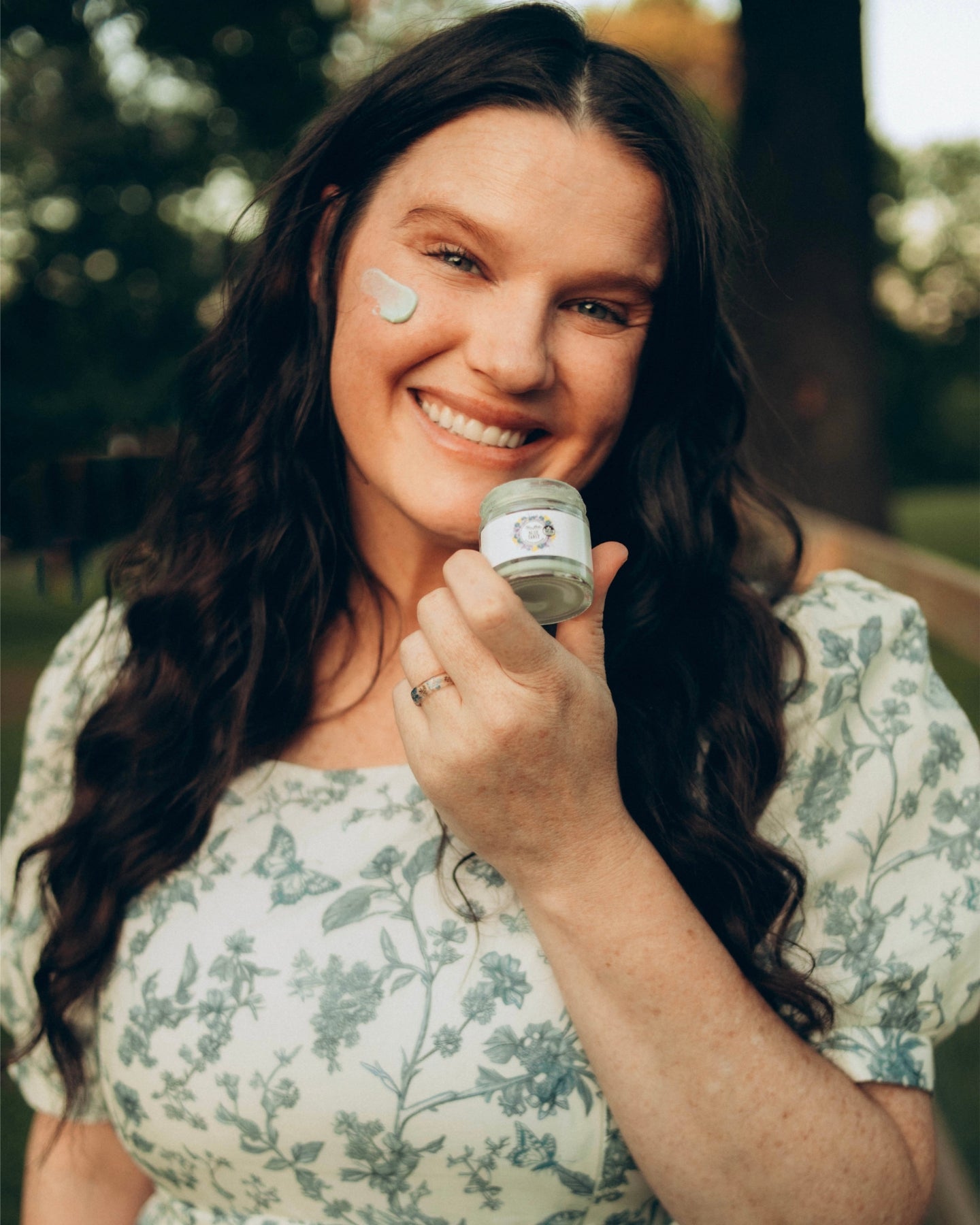 A smiling woman holds up an open jar of Blue Tansy Ultra Rich Moisturizer with a daub of the product on her cheek.