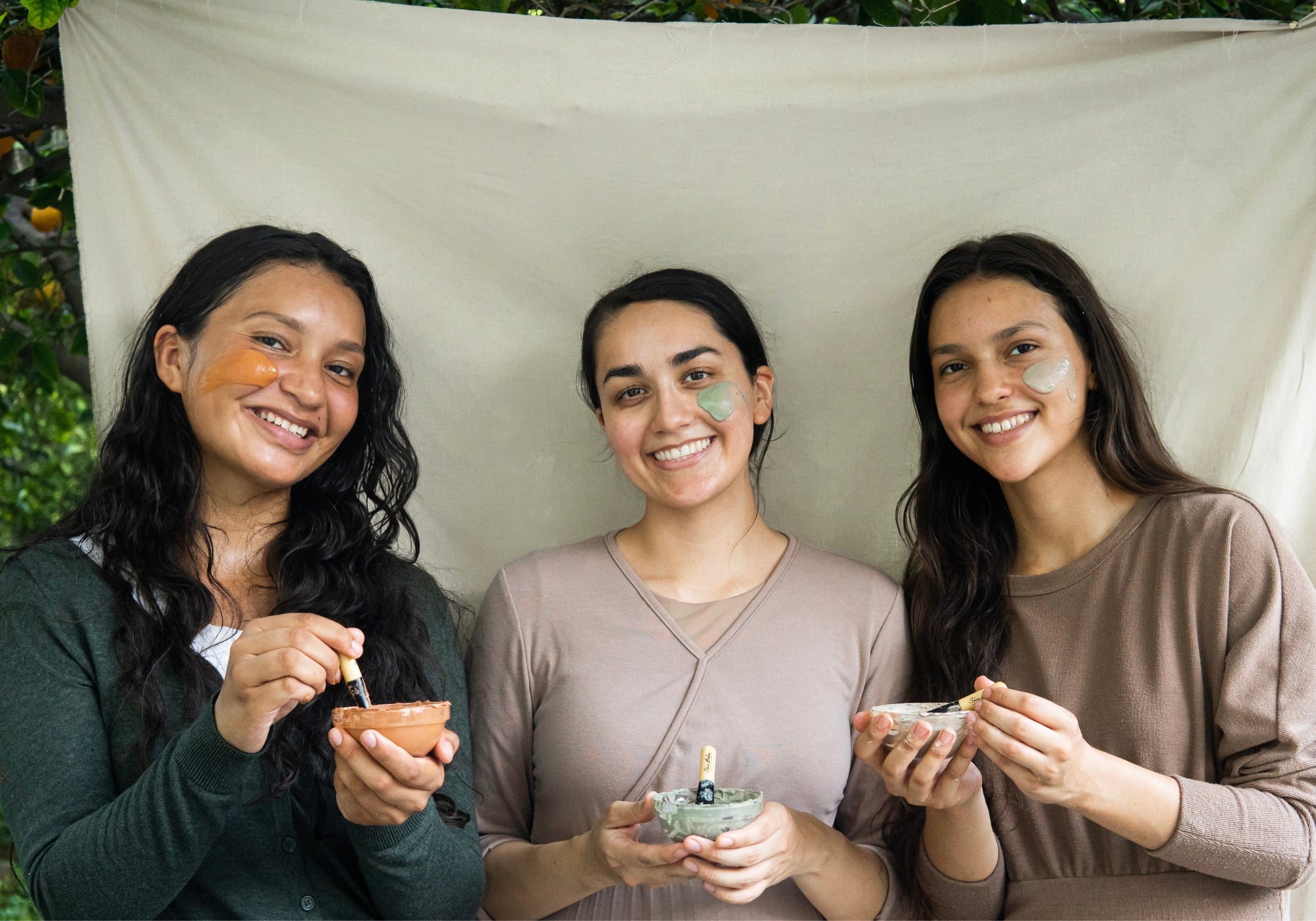 Three smiling women with skin care masks on their face in front of a beige linen canvas.